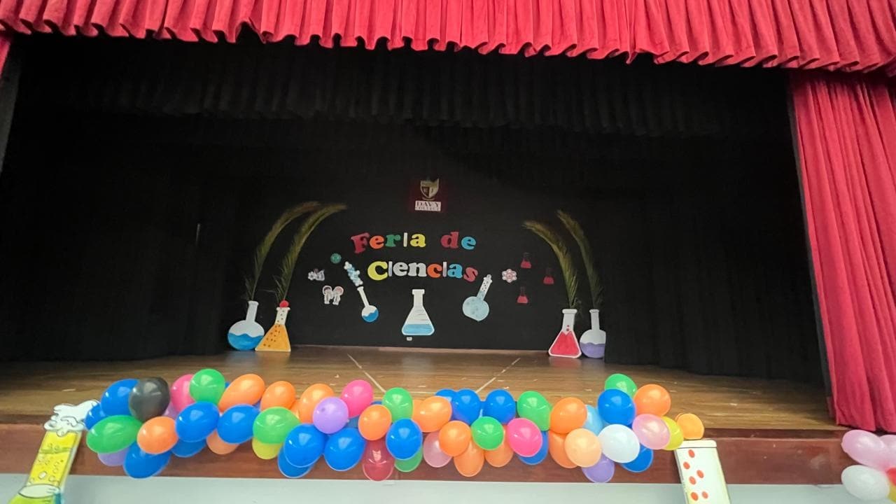Escenario del coliseo decorado con globos de colores, matraces de cartón y el letrero Feria de Ciencias