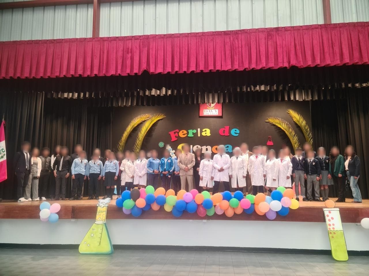 Estudiantes con mandil y uniforme de pie en el escenario decorado con el letrero Feria de Ciencias
