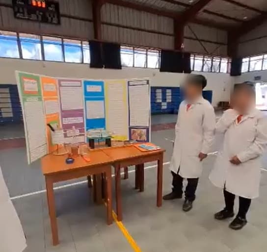 Dos estudiantes con mandil de pie junto a su panel y su prototipo