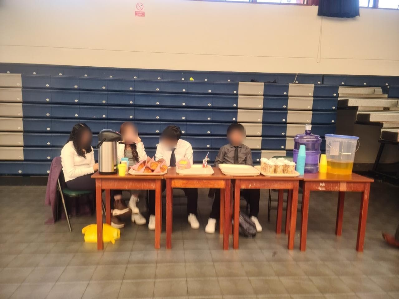 Estudiantes sentados ante una mesa con termos, jarras y bocaditos en el coliseo