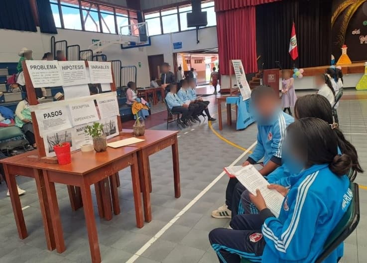 Stand con carteles de problema, hipótesis, variables y pasos; dos estudiantes repasan sus apuntes