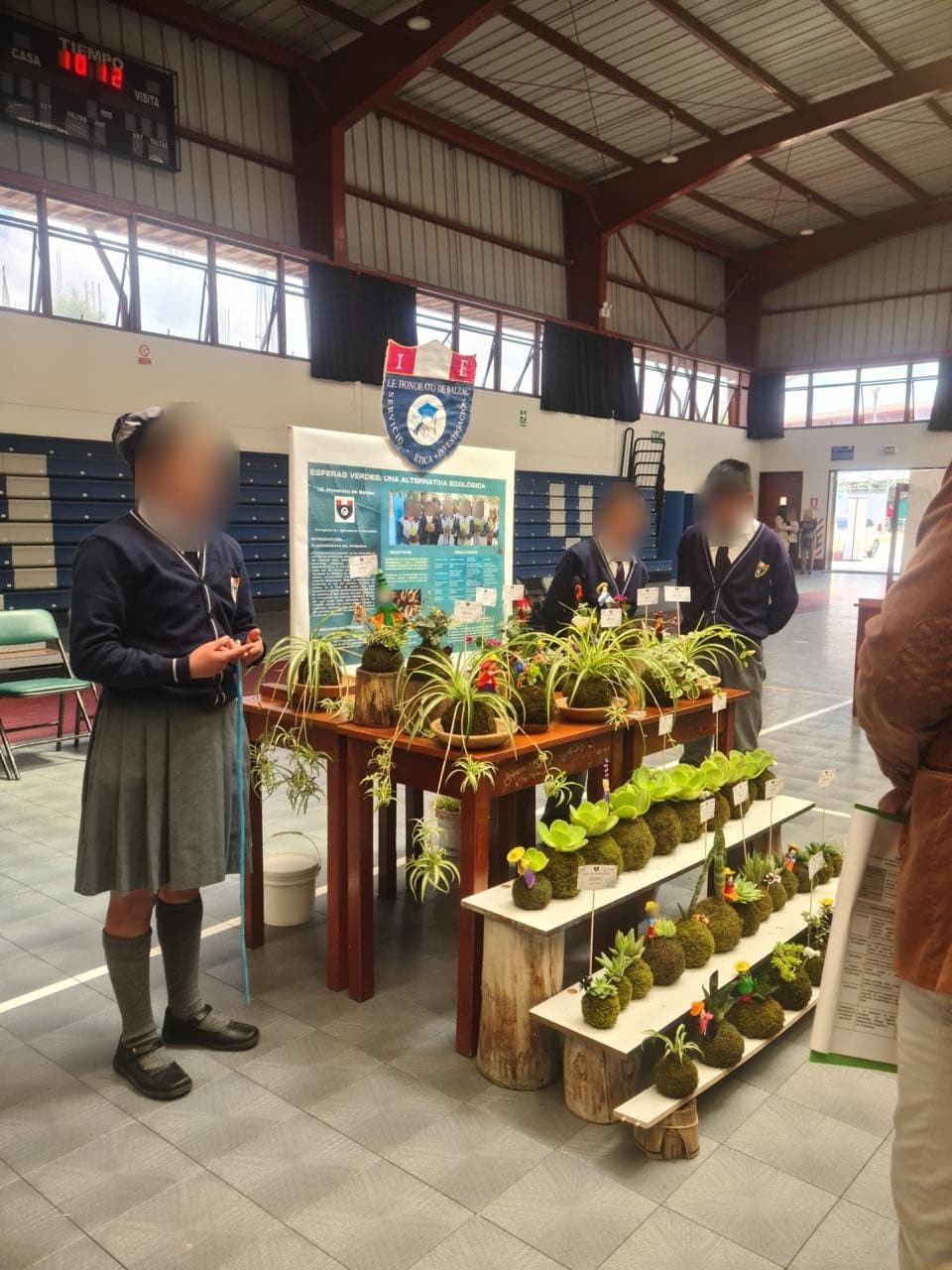 Stand del proyecto Esferas Verdes con kokedamas sobre la mesa y el estandarte de la I.E. Honorato de Balzac